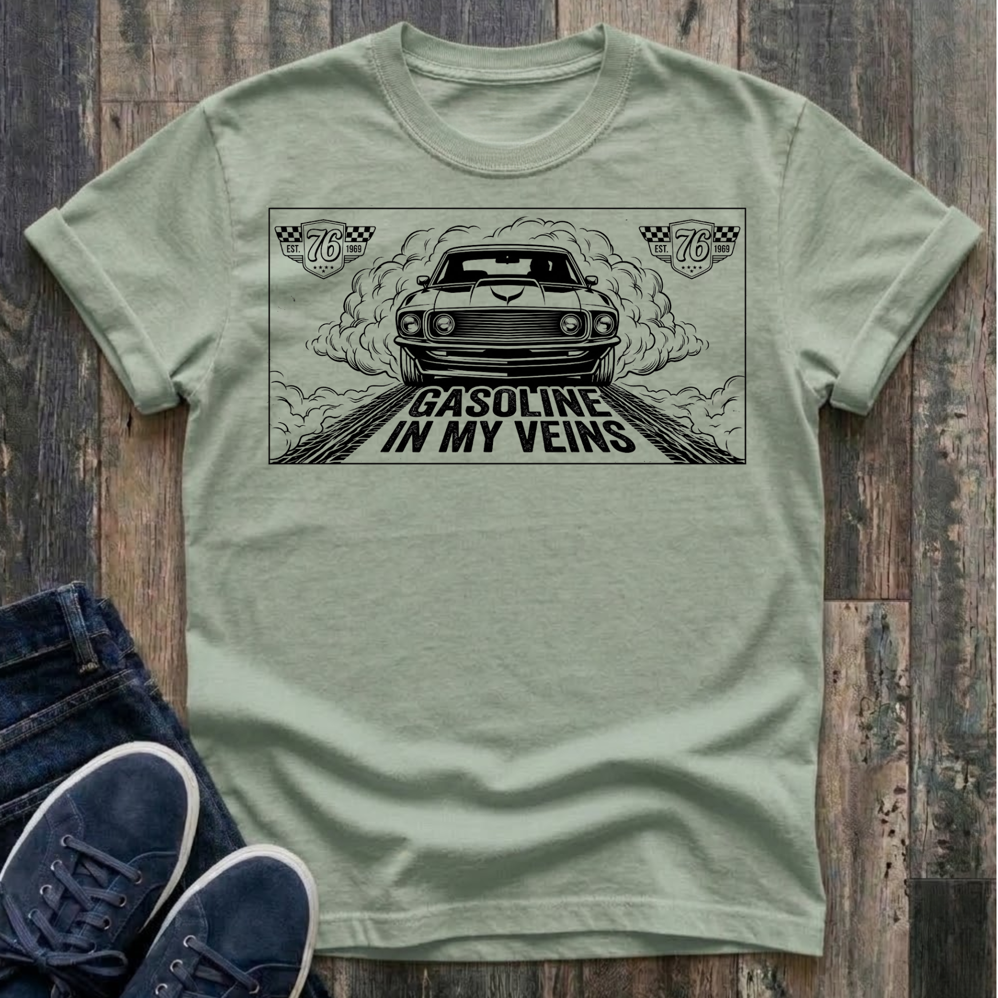 Bay t-shirt with black illustration of a classic muscle car launching forward head-on through billowing tire smoke on a drag strip, checkered racing badges reading "Est. 76 · 1969" in upper corners, bold distressed text reads "Gasoline In My Veins"
