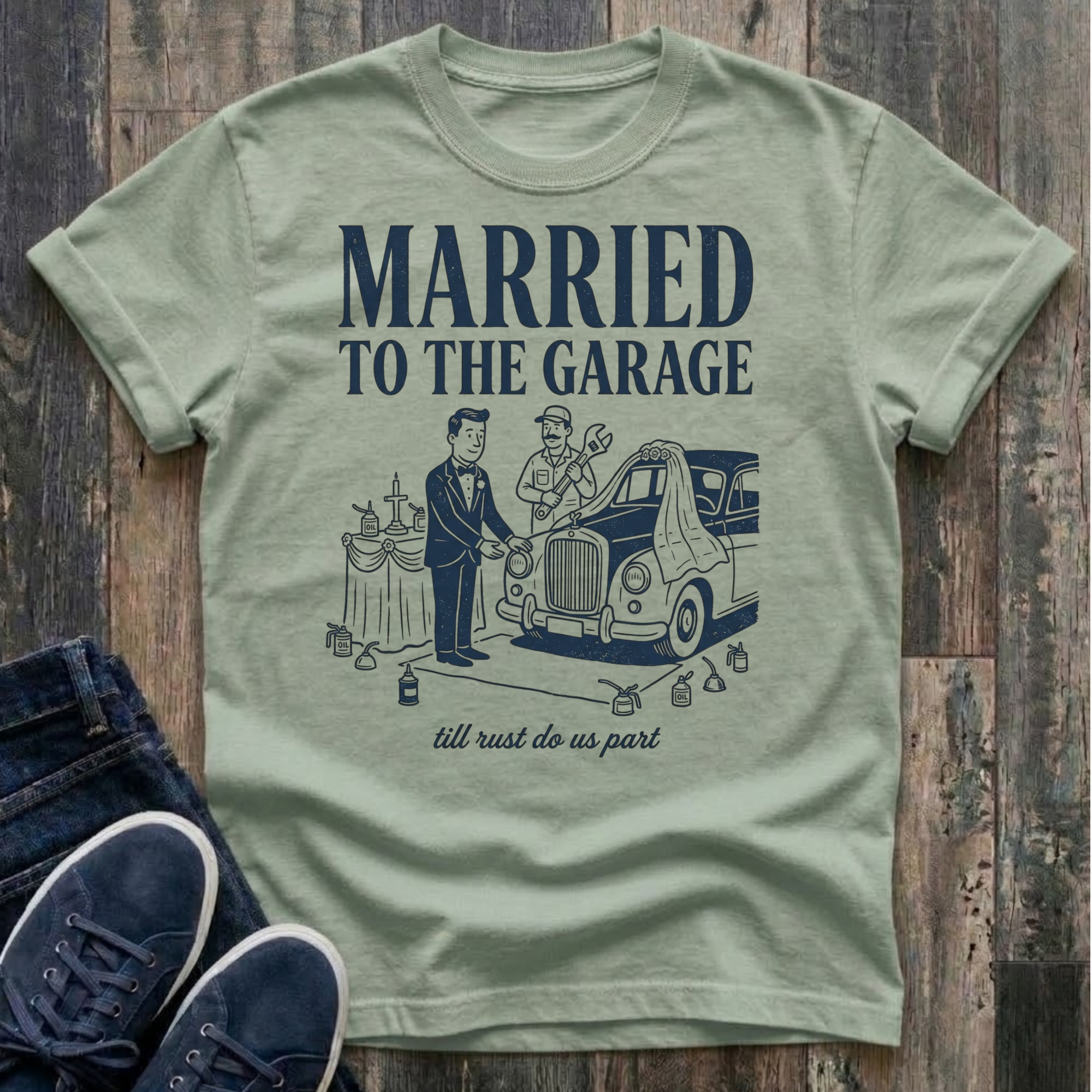 Bay vintage-style t-shirt with navy illustration of a groom in a tuxedo standing at an altar beside a classic car draped in a wedding veil, a mechanic officiating with a wrench raised, oil cans lining the aisle, text reads "Married To The Garage · till rust do us part"