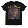A black short-sleeve graphic T-shirt on a white background, featuring a Halloween-themed illustration of a skeleton sitting in an armchair and viewing parked cars through a window, with the text 'HAUNTED BY THE CARS I DIDN'T BUY.' and decorative autumn accents.