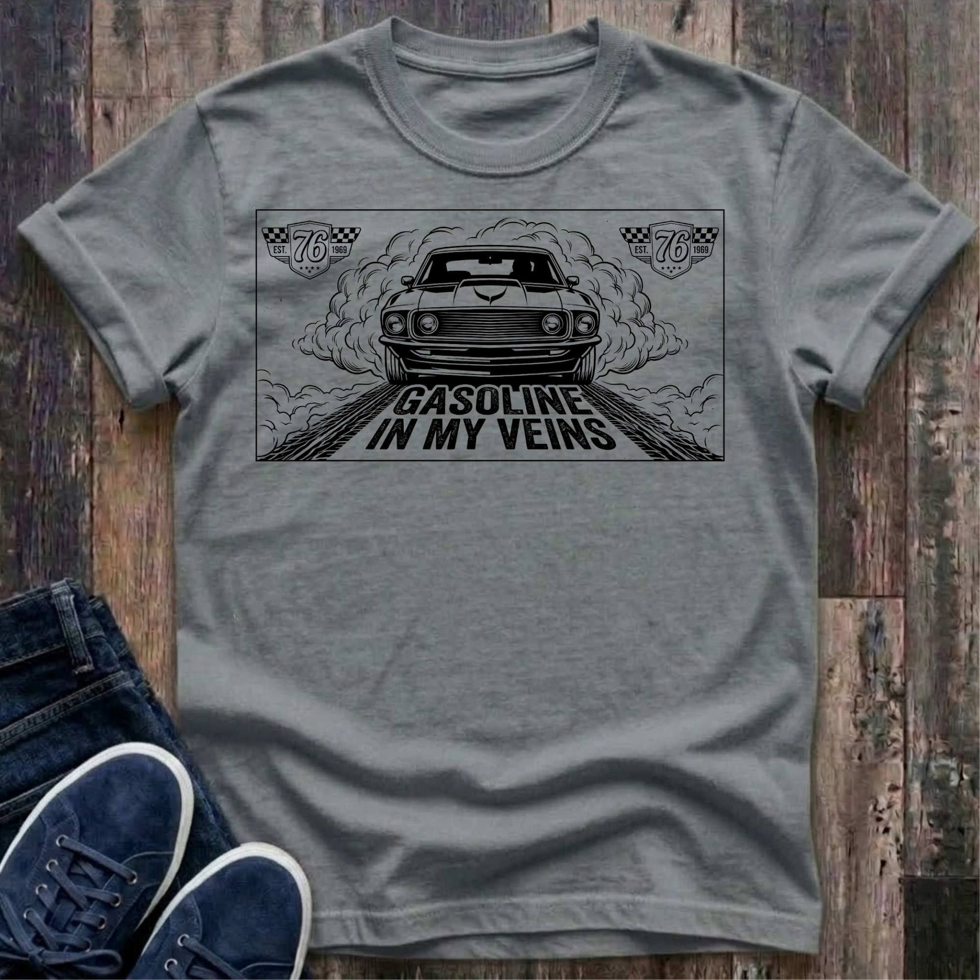 Granite t-shirt with black illustration of a classic muscle car launching forward head-on through billowing tire smoke on a drag strip, checkered racing badges reading "Est. 76 · 1969" in upper corners, bold distressed text reads "Gasoline In My Veins"
