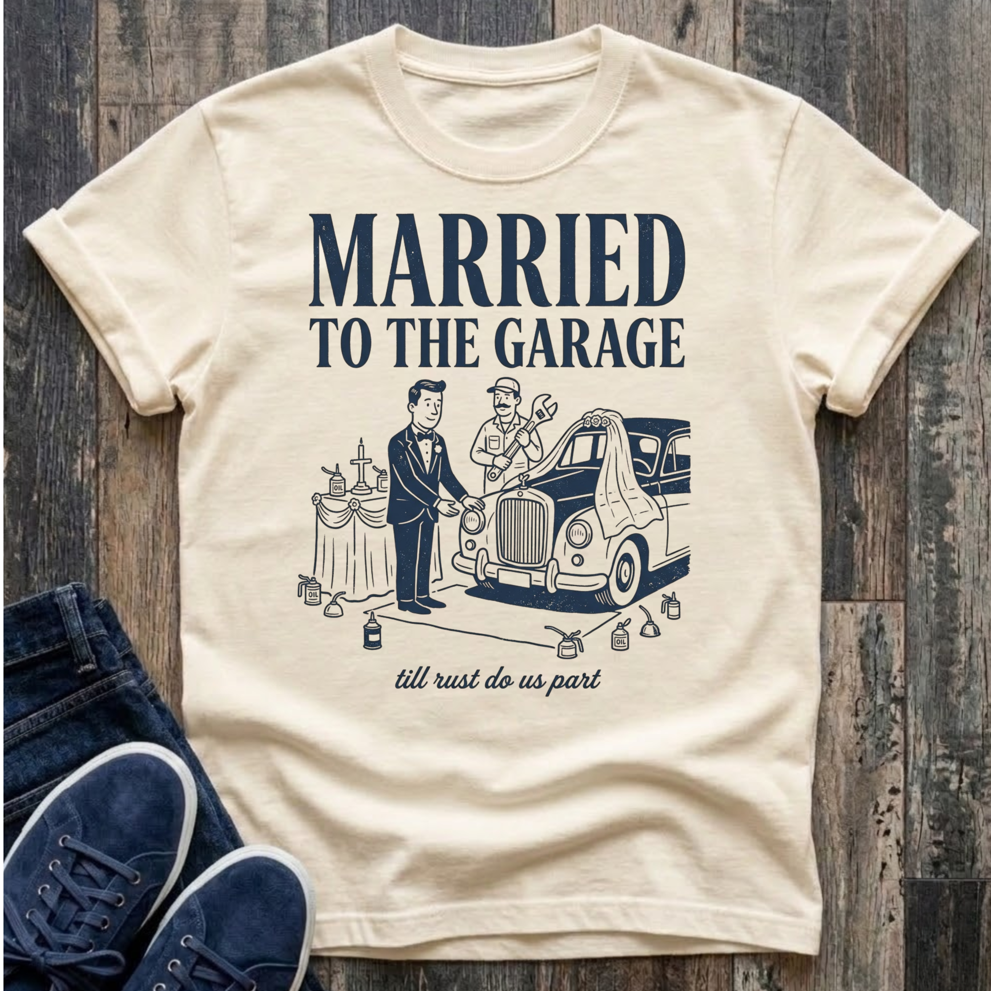 Cream vintage-style t-shirt with navy illustration of a groom in a tuxedo standing at an altar beside a classic car draped in a wedding veil, a mechanic officiating with a wrench raised, oil cans lining the aisle, text reads "Married To The Garage · till rust do us part"
