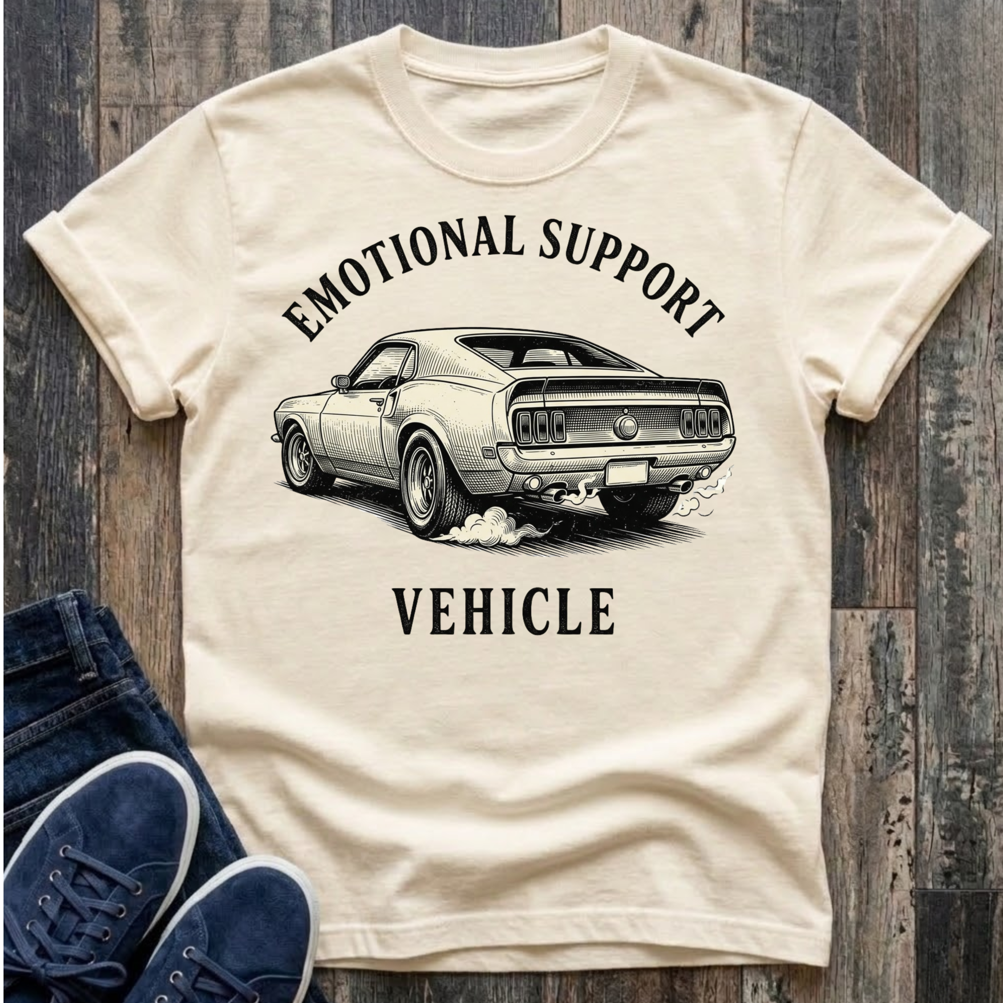 Beige t-shirt with a graphic of a car and text 'Emotional Support Vehicle' on a wooden surface.