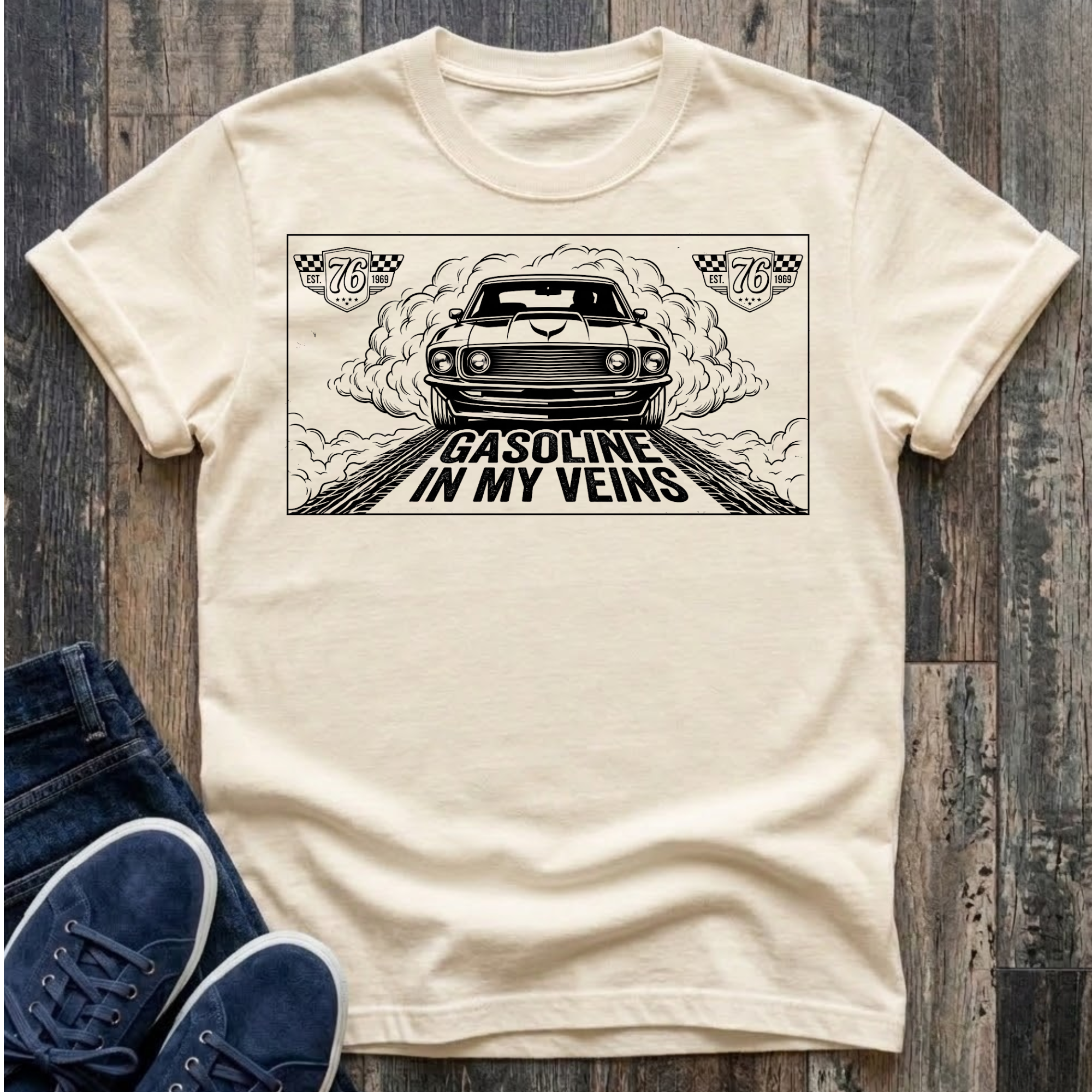 Ivory t-shirt with black illustration of a classic muscle car launching forward head-on through billowing tire smoke on a drag strip, checkered racing badges reading "Est. 76 · 1969" in upper corners, bold distressed text reads "Gasoline In My Veins"
