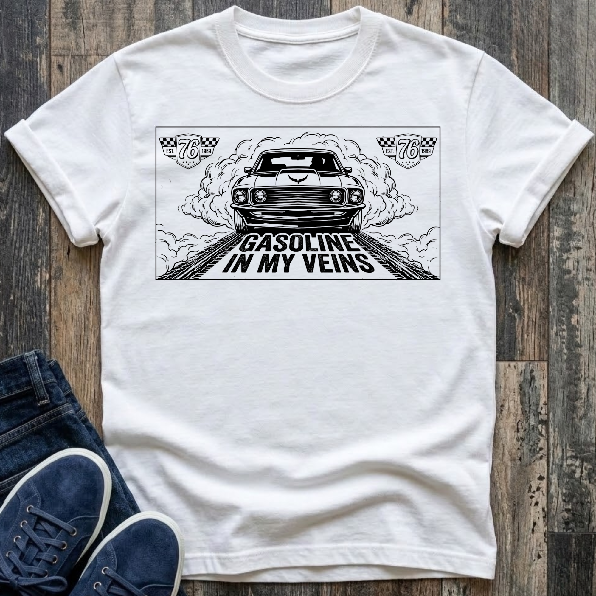 White t-shirt with black illustration of a classic muscle car launching forward head-on through billowing tire smoke on a drag strip, checkered racing badges reading "Est. 76 · 1969" in upper corners, bold distressed text reads "Gasoline In My Veins"
