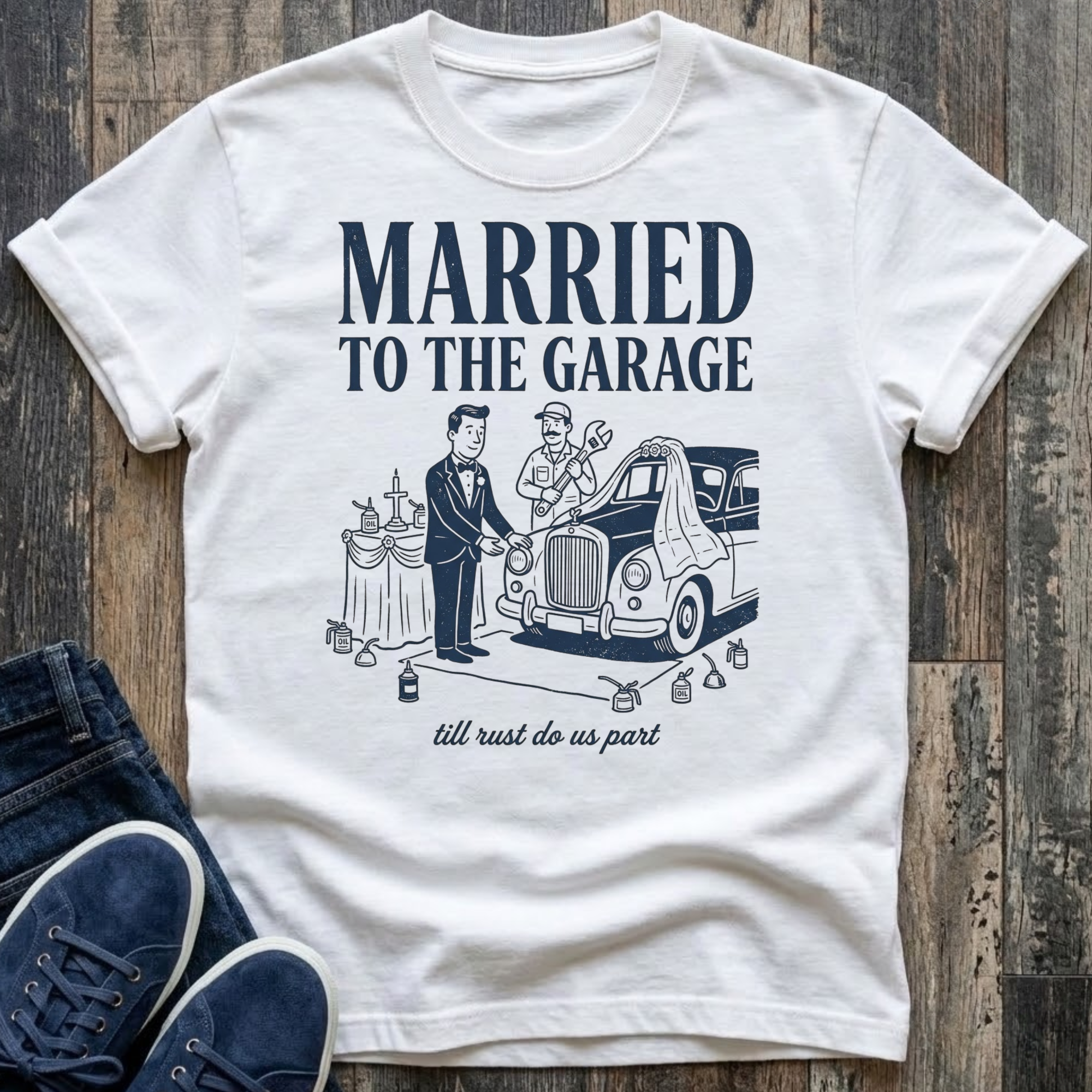 white vintage-style t-shirt with navy illustration of a groom in a tuxedo standing at an altar beside a classic car draped in a wedding veil, a mechanic officiating with a wrench raised, oil cans lining the aisle, text reads "Married To The Garage · till rust do us part"