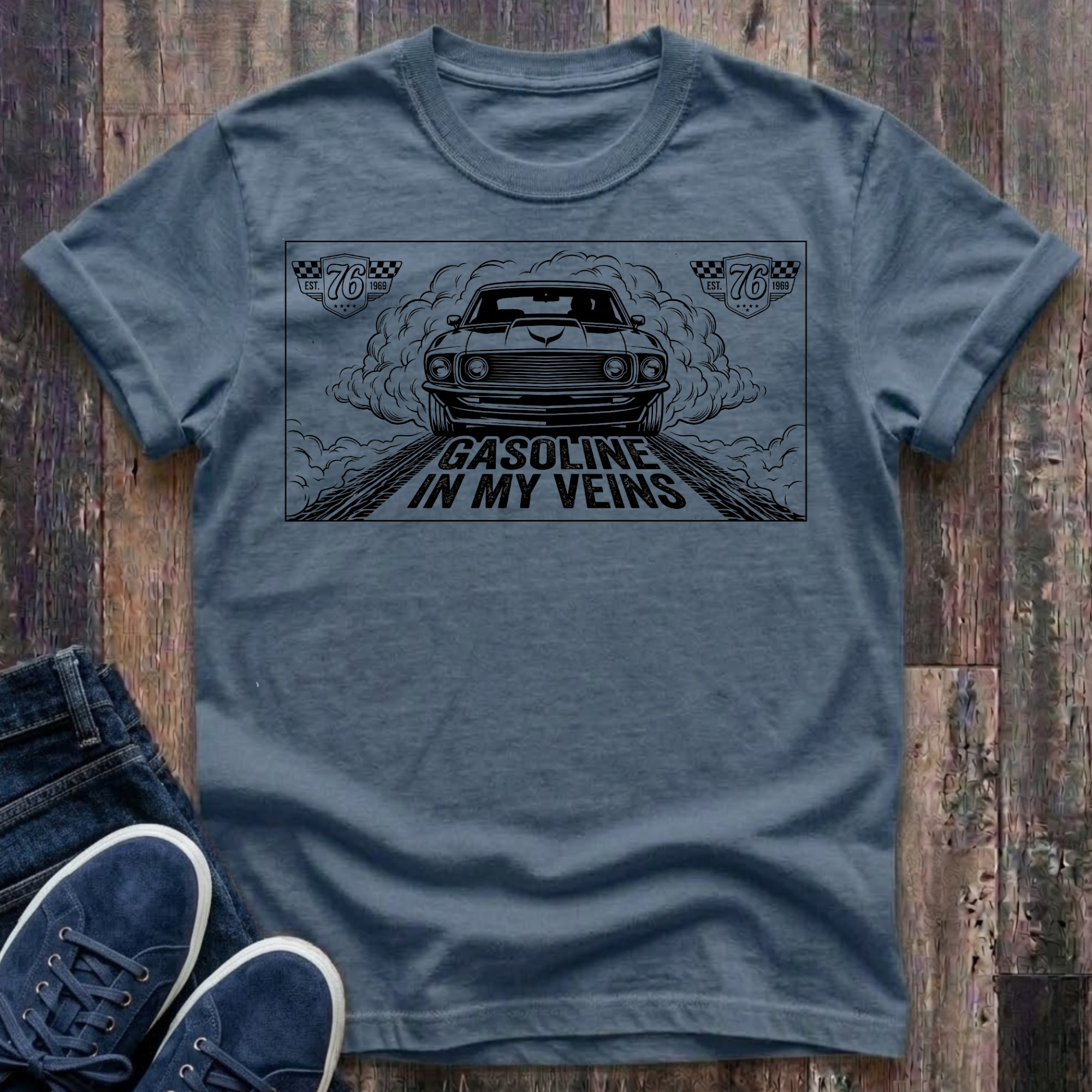 blue jean t-shirt with black illustration of a classic muscle car launching forward head-on through billowing tire smoke on a drag strip, checkered racing badges reading "Est. 76 · 1969" in upper corners, bold distressed text reads "Gasoline In My Veins"
