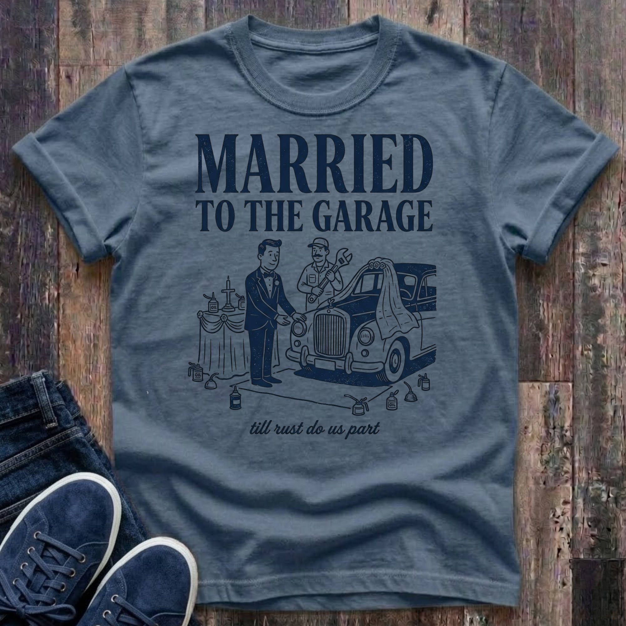 blue jean vintage-style t-shirt with navy illustration of a groom in a tuxedo standing at an altar beside a classic car draped in a wedding veil, a mechanic officiating with a wrench raised, oil cans lining the aisle, text reads "Married To The Garage · till rust do us part"