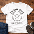 Eat Sleep Soccer Repeat T-Shirt with a soccer ball graphic, laid on a wooden surface with jeans and shoes.