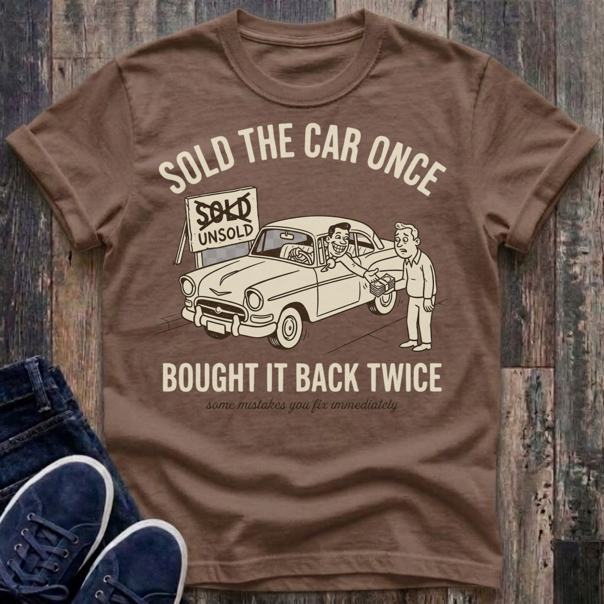 Espresso vintage-style t-shirt with cream illustration of a grinning man sitting behind the wheel of a classic car handing cash back to a confused buyer, a sign reading "SOLD" crossed out and replaced with "UNSOLD" in the background, text reads "Sold The Car Once · Bought It Back Twice · some mistakes you fix immediately"