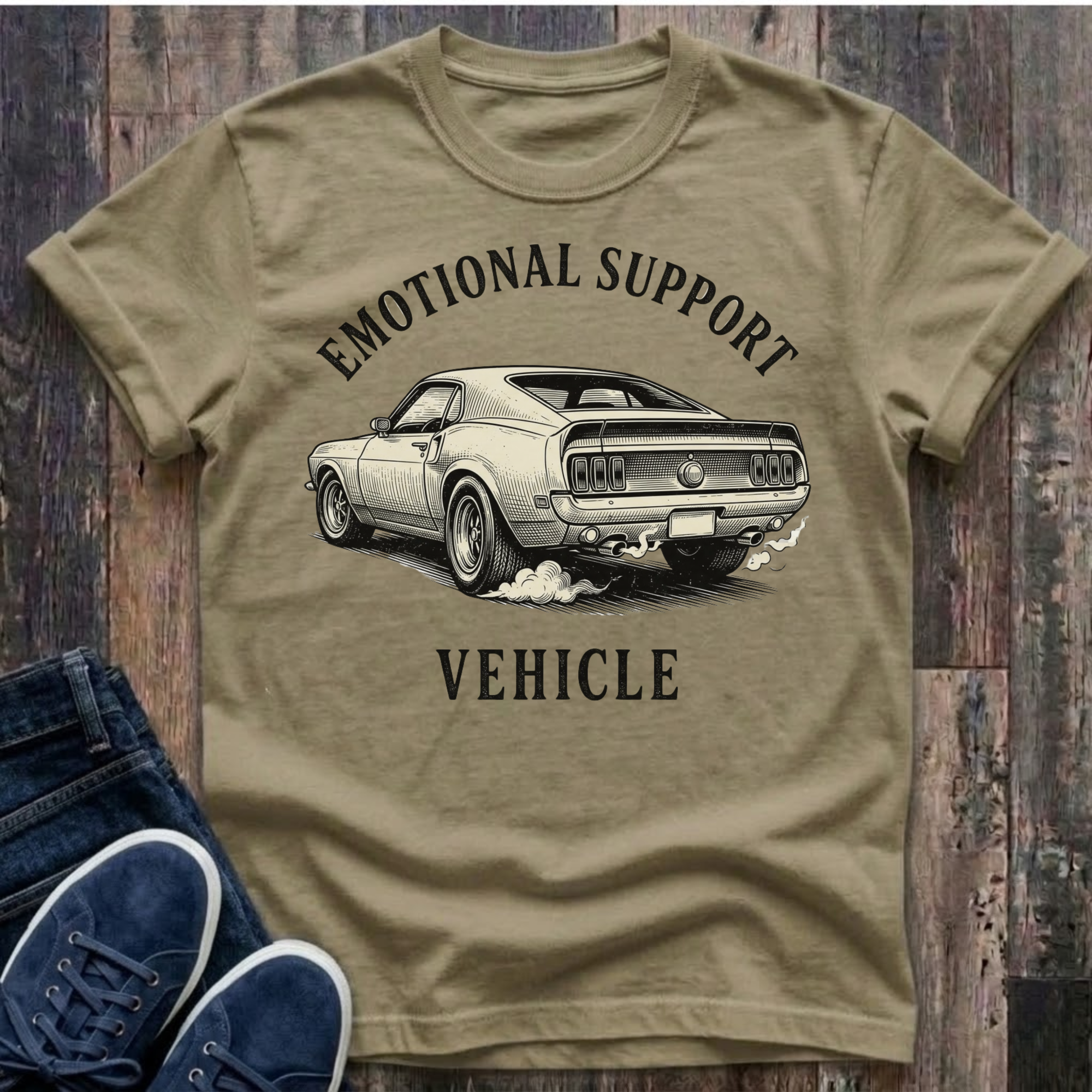 Khaki t-shirt with a graphic of a car and text 'Emotional Support Vehicle' on a wooden surface with blue sneakers.