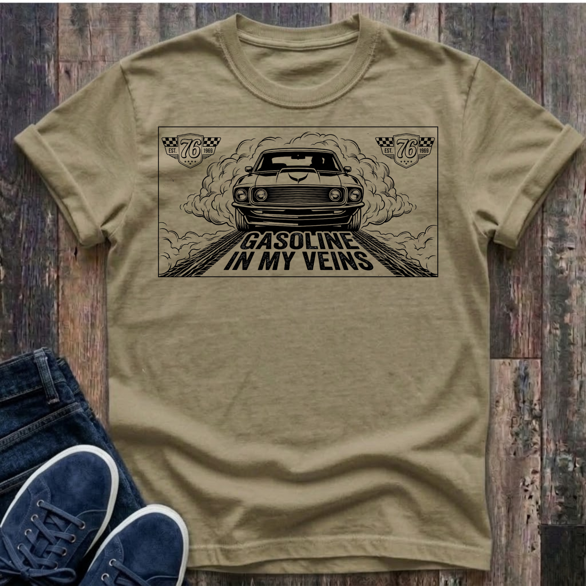khaki t-shirt with black illustration of a classic muscle car launching forward head-on through billowing tire smoke on a drag strip, checkered racing badges reading "Est. 76 · 1969" in upper corners, bold distressed text reads "Gasoline In My Veins"
