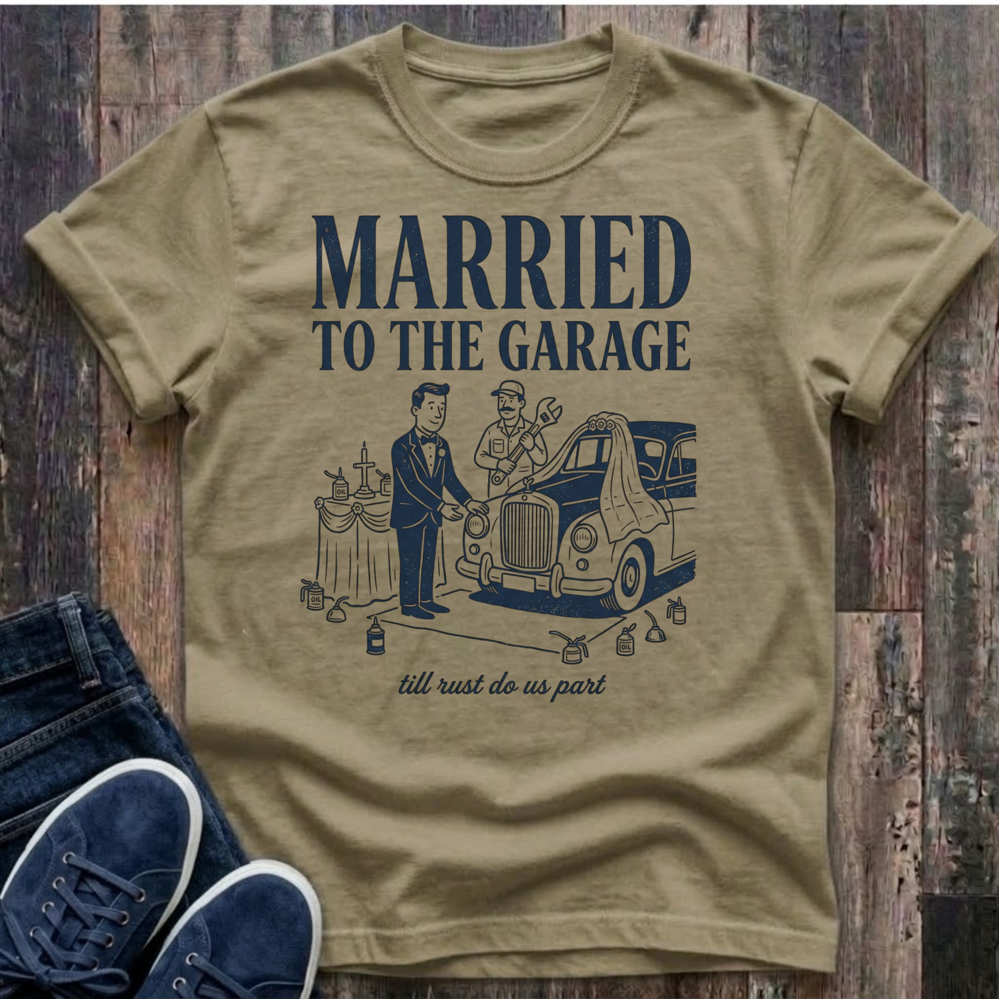 khaki vintage-style t-shirt with navy illustration of a groom in a tuxedo standing at an altar beside a classic car draped in a wedding veil, a mechanic officiating with a wrench raised, oil cans lining the aisle, text reads "Married To The Garage · till rust do us part"